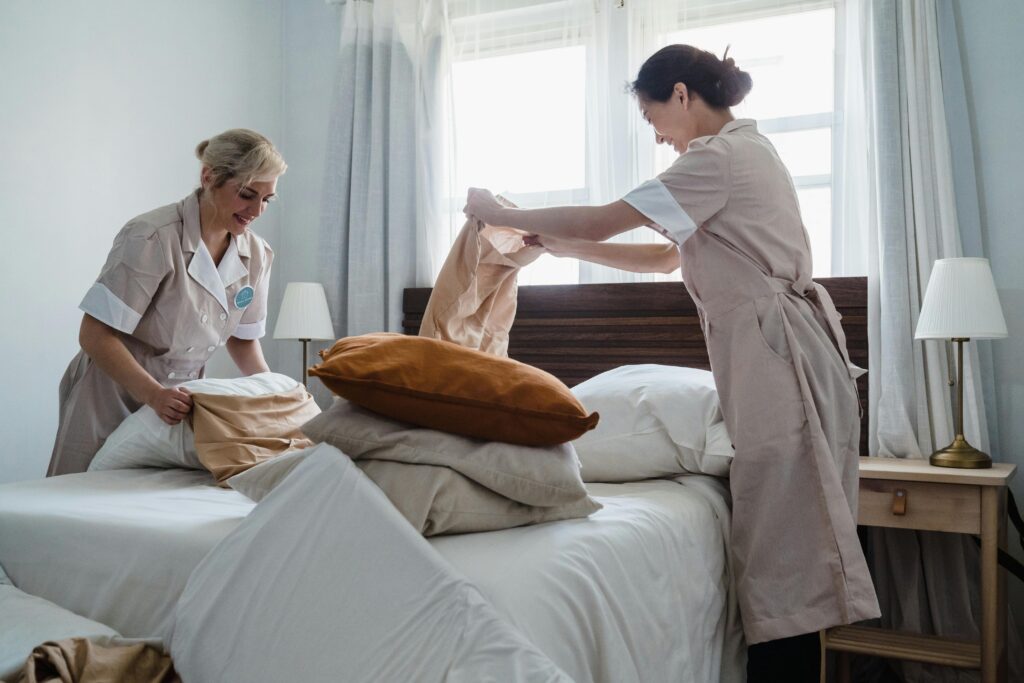 pexels photo 9462745 9462745 Two housekeepers in uniform fixing a hotel room bed with fresh linen and pillows.